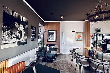 Moderner Innenraum einer Pizzeria mit Tischen, Stühlen, Wandbildern und einer Getränkekarte an der Wand. Größtenteils in grau-weiß eingerichtet mit Holzoptik-Fassaden an der Wand.