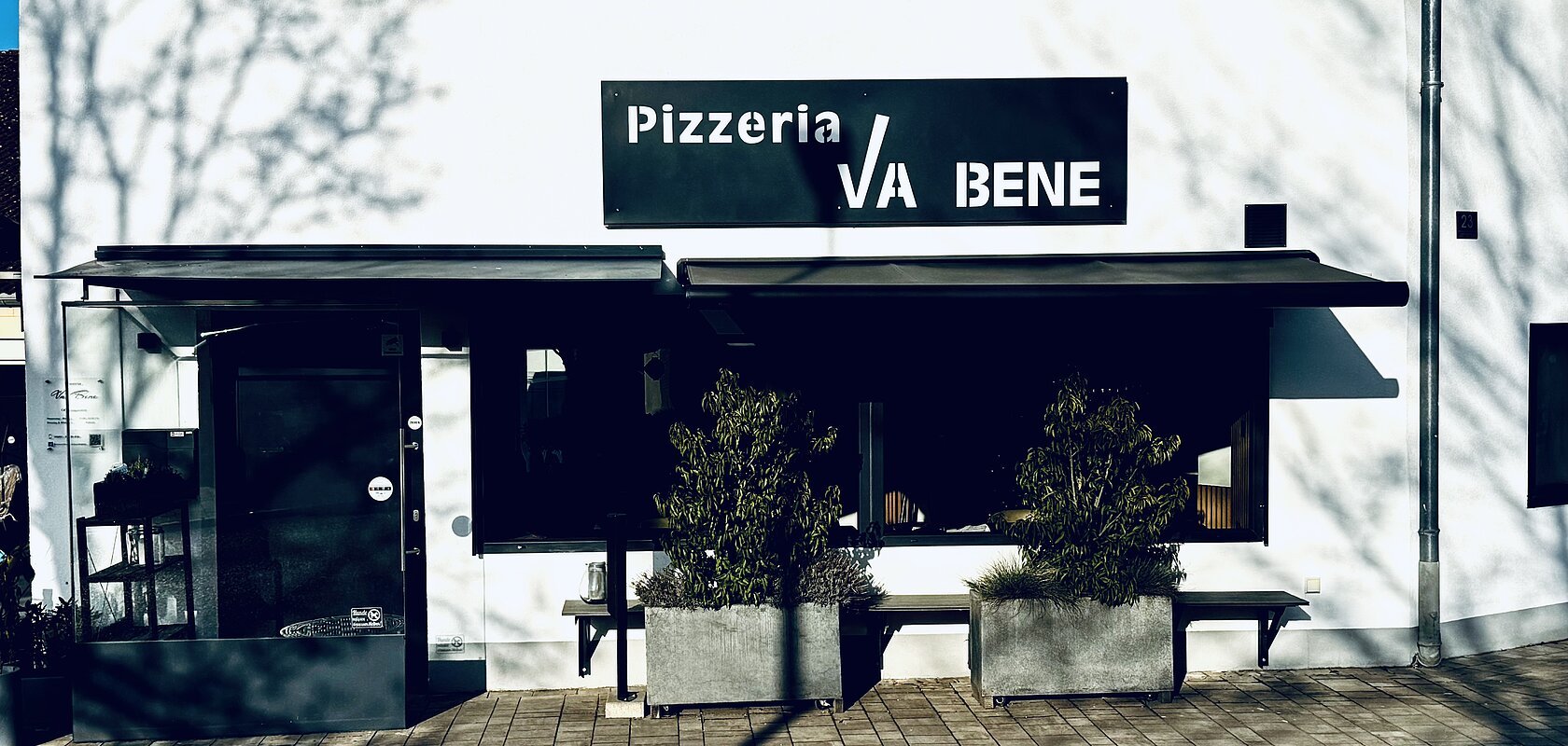 Außenansicht einer Pizzeria mit schwarzem Schild „Pizzeria VA BENE“, eine Eingansbereich aus Glas, verdunkeltem großen Fenster mit Rollläden und zwei großen Pflanzkübeln davor. Vor der Pizzeria ein gepflasterter Gehweg.