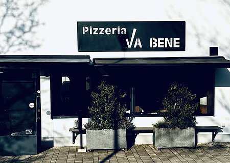 Außenansicht einer Pizzeria mit schwarzem Schild „Pizzeria VA BENE“, eine Eingansbereich aus Glas, verdunkeltem großen Fenster mit Rollläden und zwei großen Pflanzkübeln davor. Vor der Pizzeria ein gepflasterter Gehweg.