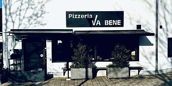 Außenansicht einer Pizzeria mit schwarzem Schild „Pizzeria VA BENE“, eine Eingansbereich aus Glas, verdunkeltem großen Fenster mit Rollläden und zwei großen Pflanzkübeln davor. Vor der Pizzeria ein gepflasterter Gehweg.