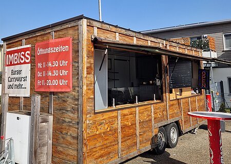 Holz-Imbisswagen mit geöffnetem Fenster, roten Schildern mit Speisen und Öffnungszeiten, stehend auf Asphalt bei blauem Himmel und einem weiteren Haus im Hintergrund.