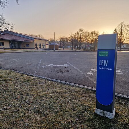 Blaue Ladestation für Elektroautos mit der Aufschrift LEW, auf einem leeren gepflastertem Parkplatz vor einem orangem Gebäude im Hintergrund bei Sonnenaufgang.