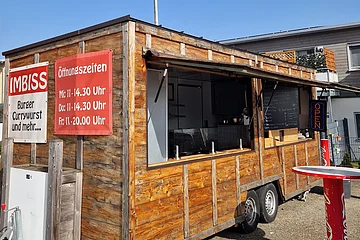 Holz-Imbisswagen mit geöffnetem Fenster, roten Schildern mit Speisen und Öffnungszeiten, stehend auf Asphalt bei blauem Himmel und einem weiteren Haus im Hintergrund.