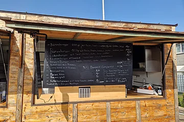 Foto einer schwarzen Speisekarte mit Burgern, Pommes, Currywurst und Preisen an einem Holzimbiss bei Sonnenschein.