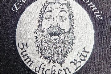 Logo mit einem Foto eines lachendem Mannes mit langem Bart und Brille. Darüber der Schriftzug „Eventgastronomie und unterhalb die Schrift "Zum dicken Bär“ auf dunklem Stoff.