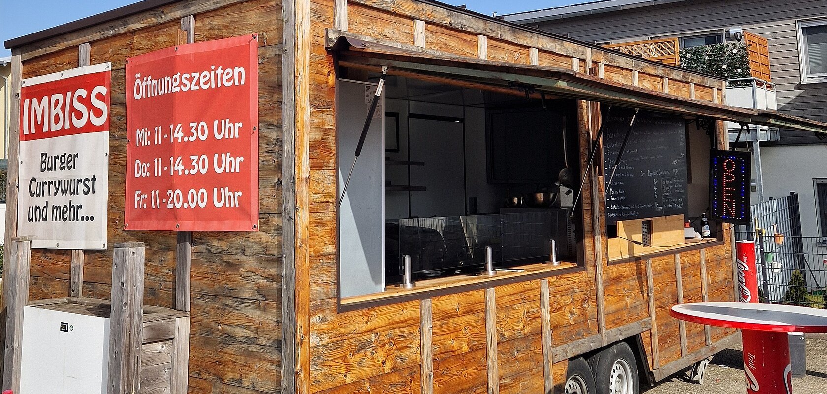 Holz-Imbisswagen mit geöffnetem Fenster, roten Schildern mit Speisen und Öffnungszeiten, stehend auf Asphalt bei blauem Himmel und einem weiteren Haus im Hintergrund.
