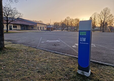 Blaue Ladestation für Elektroautos mit der Aufschrift LEW, auf einem leeren gepflastertem Parkplatz vor einem orangem Gebäude im Hintergrund bei Sonnenaufgang.