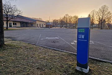 Blaue Ladestation für Elektroautos mit der Aufschrift LEW, auf einem leeren gepflastertem Parkplatz vor einem orangem Gebäude im Hintergrund bei Sonnenaufgang.