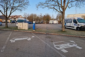 Parkplätze mit Symbolen für Elektroautos auf dem Asphalt und einer dahinterstehender Ladestation in blau, im Freien bei Tageslicht. Im Hintergrund ein Wohnmobilstellplatz mit Parkflächen und mit verschiedenen Fahrzeugen drauf.