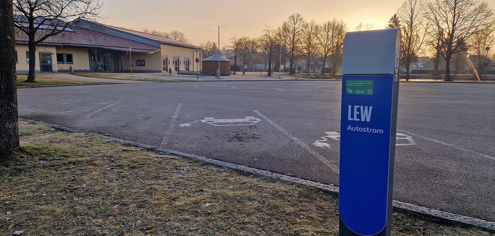 Blaue Ladestation für Elektroautos mit der Aufschrift LEW, auf einem leeren gepflastertem Parkplatz vor einem orangem Gebäude im Hintergrund bei Sonnenaufgang.