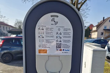 Ladesäule für Elektrofahrzeuge stehend auf einer Grünfläche bei Tageslicht, mit Bedienungsanleitung und Förderhinweis des Bundesministeriums für Verkehr in Wohngebiet.
