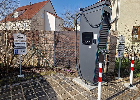 Schwarze Elektroauto-Ladestation mit zwei Ladepunkten stehend vor einem Parkplatz mit Schildern zur Parkzeitbegrenzung auf 2 Stunden, bei Tageslicht. Im Hintergrund Wohnhäuser vor blauem Himmel.