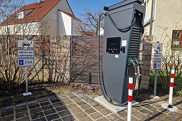Schwarze Elektroauto-Ladestation mit zwei Ladepunkten stehend vor einem Parkplatz mit Schildern zur Parkzeitbegrenzung auf 2 Stunden, bei Tageslicht. Im Hintergrund Wohnhäuser vor blauem Himmel.