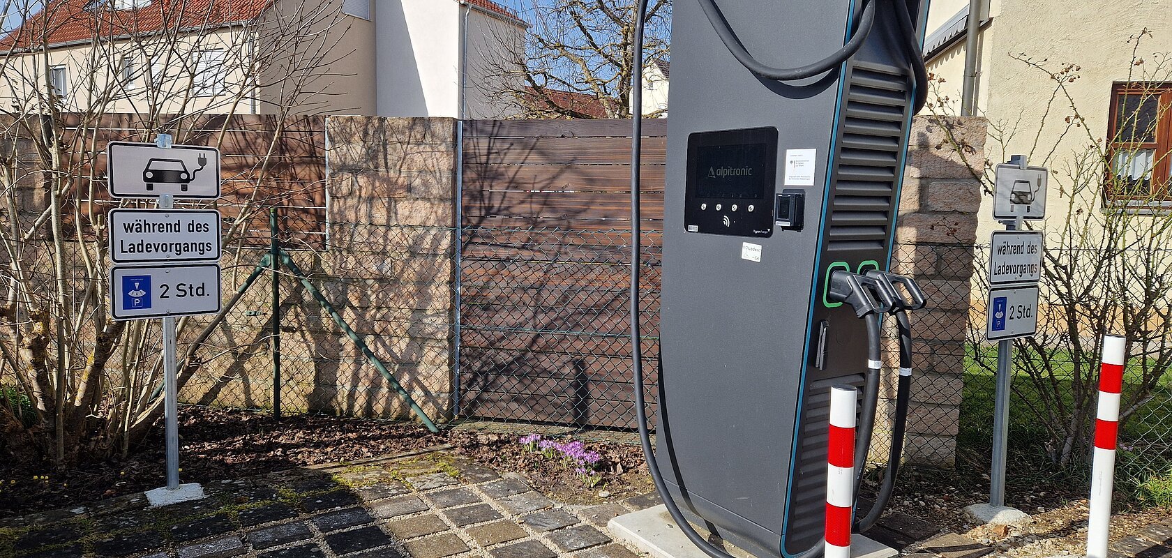Schwarze Elektroauto-Ladestation mit zwei Ladepunkten stehend vor einem Parkplatz mit Schildern zur Parkzeitbegrenzung auf 2 Stunden, bei Tageslicht. Im Hintergrund Wohnhäuser vor blauem Himmel.