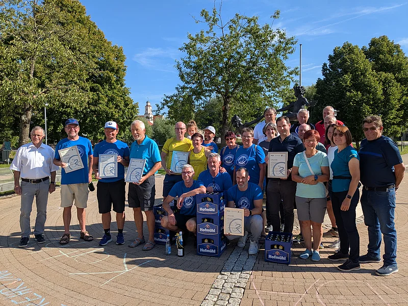Gruppenfoto von Personen, die im Freien auf einem gepflastertem Platz stehen, einige halten Urkunden in den Händen. Neben der Gruppe stehen vier blaue Getränkekisten. Im Hintergrund sind Bäume, blauer Himmel, ein Kirchenturm und eine Statue, bei Sonnenschein.