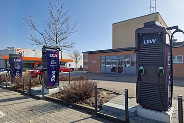 Drei dunkle Ladestationen für Elektroautos an Parkplätzen mit jeweils zwei Ladepunkten. Im Hintergrund ein oranges Gebäude, der Drogeriemarkt Müller und mehrere parkende Autos daneben.