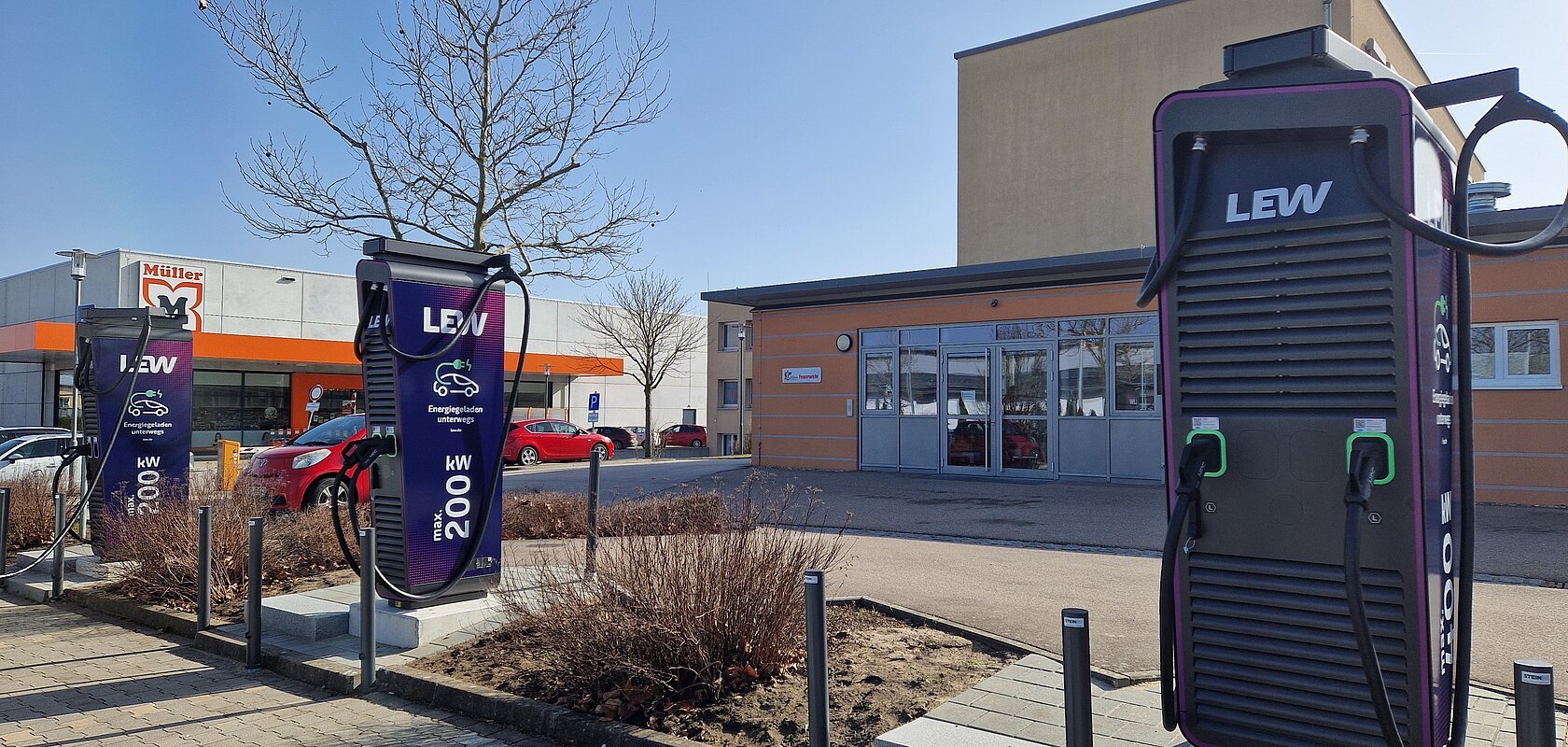 Drei dunkle Ladestationen für Elektroautos an Parkplätzen mit jeweils zwei Ladepunkten. Im Hintergrund ein oranges Gebäude, der Drogeriemarkt Müller und mehrere parkende Autos daneben.