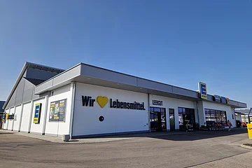 Außenansicht eines Edeka-Supermarkts auf einem asphaltiertem Platz mit Schriftzug „Wir lieben Lebensmittel“ an der weißen Fassade und klarem Himmel im Hintergrund.