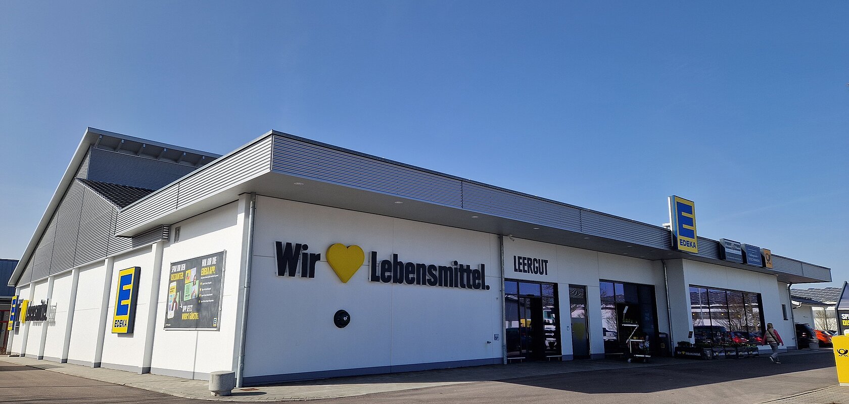 Außenansicht eines Edeka-Supermarkts auf einem asphaltiertem Platz mit Schriftzug „Wir lieben Lebensmittel“ an der weißen Fassade und klarem Himmel im Hintergrund.