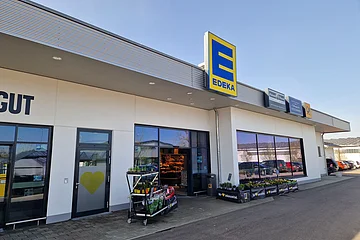 Eingang eines Edeka-Supermarkts mit Pflanzenverkauf und Parkplätzen im Hintergrund bei klarem Himmel.
