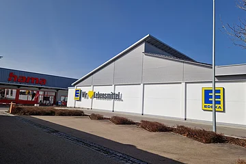 Außenansicht eines Edeka-Supermarkts mit Schriftzug „Wir lieben Lebensmittel“ an der weißen Fassade und links daneben ein Hama-Geschäft. Im Vordergrund ein gepflasterter Platz.