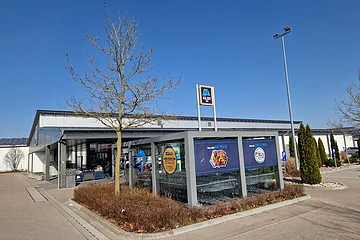 Außenansicht eines Aldi Süd Supermarkts mit Einkaufswagenunterstand, großem umgebenden Parkplatz und kahlem Bäumen bei klarem Himmel.