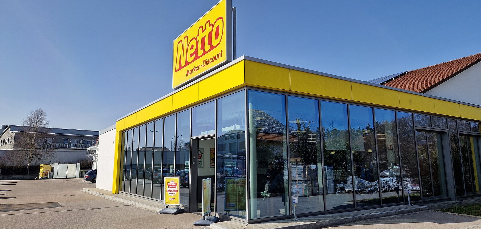 Gebäude eines Netto Marken-Discounts mit einer Glasfassade, gelbem Fassadenband und dem Schriftzug "Netto" oben auf dem Gebäude in rot/gelb. Großer Parkplatz bei klarem Himmel.