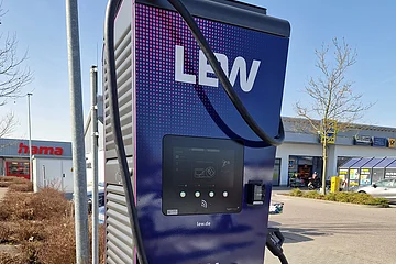 Dunkle Ladestation für Elektroautos mit Anzeige auf Display und Kabeln zum Aufladen, im Freien auf einem Parkplatz vor Einkaufsgeschäften, bei Sonnenschein.