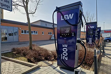 Drei dunkle Elektroauto-Ladestationen mit zwei Ladesäulen die je 200 kW Leistung bieten und einer Ladesäule mit 400 kW Leistung, an einem Parkplatz vor einem orangefarbenen Gebäude mit Flachdach.