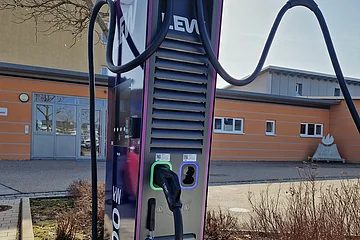 Ladesäule für Elektrofahrzeuge mit zwei Ladekabeln vor einem orangefarbenen Gebäude im Freien, umgeben von einem kahlen Planzenbeet.