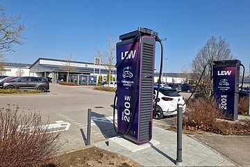 Zwei Ladestationen für Elektroautos mit 200 kW Leistung auf einem Parkplatz mit parkenden Autos vor einem Aldi-Markt bei klarem blauem Himmel.