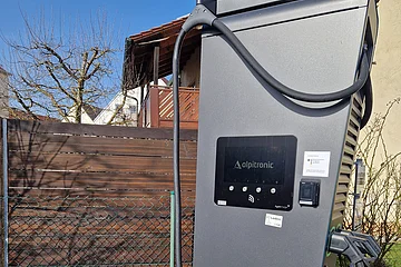 Schwarze Elektroauto-Ladesäule der Marke Alpitronic mit einem Display und zwei Schläuchen zum Laden, vor einem Holzzaun und einem Wohnhaus im Hintergrund, bei Sonnenschein.