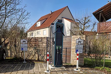 Schwarze Elektroauto-Ladestation mit zwei Ladepunkten hinter zwei gepflasterten Parkplätzen vor einem Wohnhaus bei klarem Himmel.
