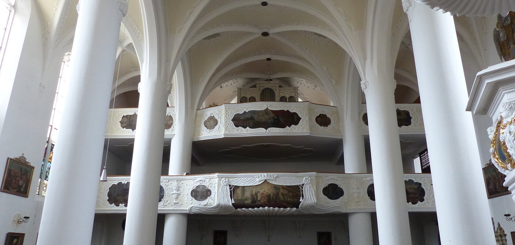 Innenansicht einer Kirche, der Stadtpfarrkirche St. Walburga in Monheim, mit hohen weißen Säulen, Gewölbedecke, Emporen mit Verzierungen und Holzbänken.