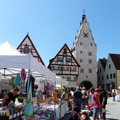 Markttreiben mit Marktständen und Menschen auf einem gepflasterten Altstadt-Platz vor Fachwerkhäusern und einem Turm mit einer Uhr, bei klarem Himmel.