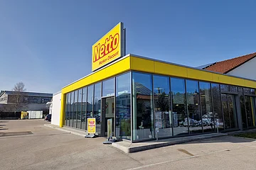 Gebäude eines Netto Marken-Discounts mit einer Glasfassade, gelbem Fassadenband und dem Schriftzug "Netto" oben auf dem Gebäude in rot/gelb. Großer Parkplatz bei klarem Himmel.