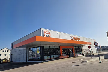 Außenansicht eines Müller-Marktes mit orangefarbenem Vordach und klarem Himmel im Hintergrund auf einem gepflastertem Platz mit Parkplätzen außenrum.