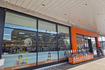 Außenansicht eines Drogeriemarktes mit großen Fenstern und orangefarbenem Eingangsbereich, Verkaufsstände vor dem Eingang, bei Tageslicht, auf einem gepflastertem Platz.