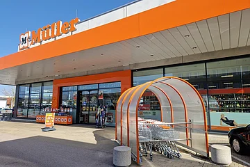 Eingangsbereich eines Müller-Marktes mit Einkaufswagen und orangen Markenzeichen "Müller" bei Sonnenschein.