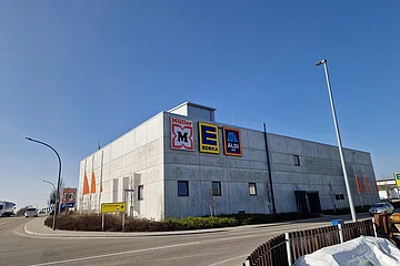 Großes graues Gebäude mit Logos von Müller, Edeka und Aldi Süd an der Fassade bei klarem Himmel, umgeben von zwei Straßen.