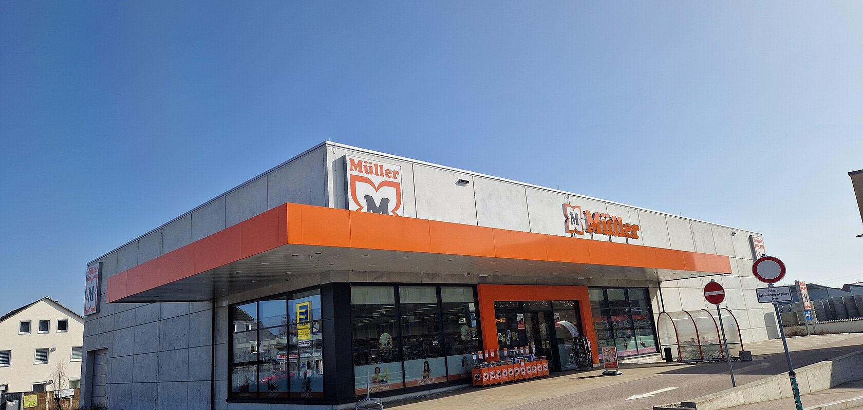 Außenansicht eines Müller-Marktes mit orangefarbenem Vordach und klarem Himmel im Hintergrund auf einem gepflastertem Platz mit Parkplätzen außenrum.