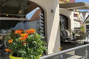 Blumentopf mit orangefarbenen Blumen auf dem Geländer einer Terrasse vor einem Café mit grauen Sitzgelegenheiten und Sonnenschirm. Auf der Fensterscheibe steht der Text "Aroma".