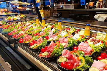 Auslage mit verschiedenen belegten Tellern, gefüllt mit Aufschnitt, Käse, Schicken, Wurst, Gemüse und Orangensaftflaschen. Im Hintergrund Kaffeemaschinen zu sehen.