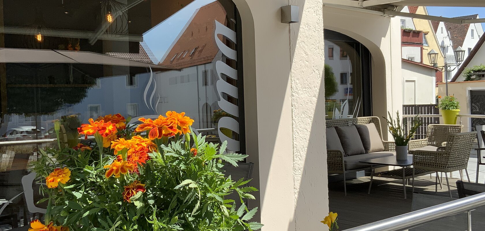 Blumentopf mit orangefarbenen Blumen auf dem Geländer einer Terrasse vor einem Café mit grauen Sitzgelegenheiten und Sonnenschirm. Auf der Fensterscheibe steht der Text "Aroma".