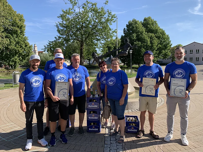 Acht Personen in blauen T-Shirts mit Urkunden und Getränkekisten vor einer Brunnen-Skulptur im Freien auf einem gepflasterten Platz. Im Hintergrund sind Bäume und blauer Himmel zu sehen.