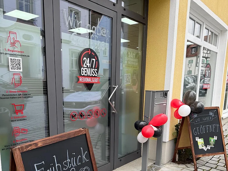 Außenansicht vom Genussmarkt 24/7 in Monheim - Regional und Gut Eingang eines 24/7 Genuss Geschäfts mit zwei schwarzen beschriebenen Kreidetafeln vor der Tür und roten, weißen und schwarzen Luftballons an den Tafeln.