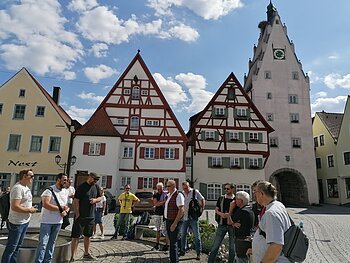 Gruppe von Menschen steht auf einem gepflasterten Platz, der Monheimer Innenstadt, vor Fachwerkhäusern und einem Turm mit Uhr. Auf einem gelben Haus davon steht der Schriftzug Nest.