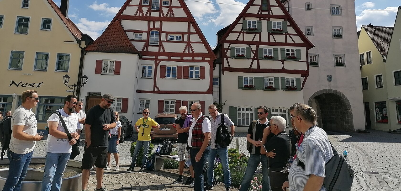 Gruppe von Menschen steht auf einem gepflasterten Platz, der Monheimer Innenstadt, vor Fachwerkhäusern und einem Turm mit Uhr. Auf einem gelben Haus davon steht der Schriftzug Nest.