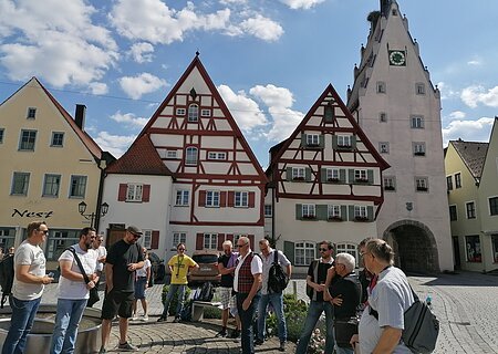 Gruppe von Menschen steht auf einem gepflasterten Platz, der Monheimer Innenstadt, vor Fachwerkhäusern und einem Turm mit Uhr. Auf einem gelben Haus davon steht der Schriftzug Nest.
