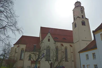 Kirchengebäude mit rotem Dach, einem Turm rechts im Bild mit Uhr und kahlen Bäumen mit grüner Wiese im Vordergrund bei bewölktem Himmel.